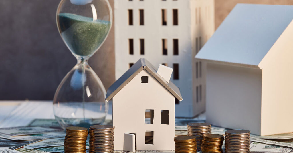 Auf einem Tisch zwischen Münzstapeln und Papiergeld stehen Hausmodelle und um Hintergrund läuft eine Sanduhr, die den Zeitdruck beim Immobiliennotverkauf symbolisiert | Notverkauf